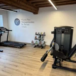 Physio Plus Geräteraum mit Schriftzug an der Wand. Kettelbell-Ständer, Laufband und  diversen Geräten zum Trainieren 