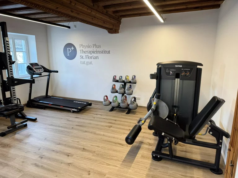 Physio Plus Geräteraum mit Schriftzug an der Wand. Kettelbell-Ständer, Laufband und  diversen Geräten zum Trainieren 